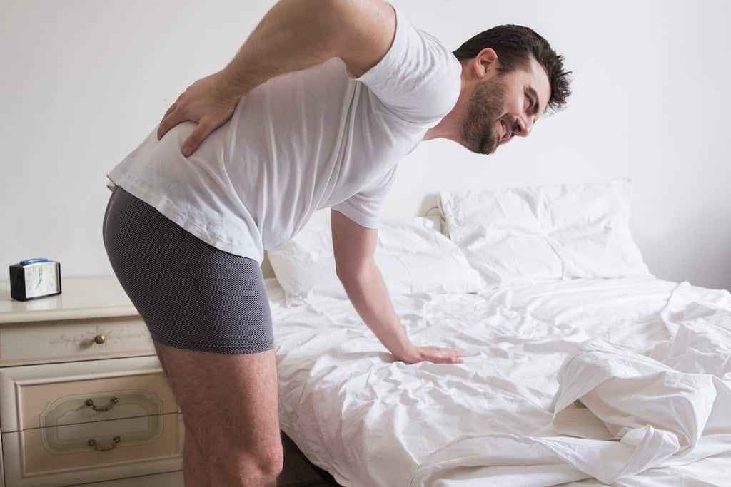 Man leaning over his bed with a hand over his lower back